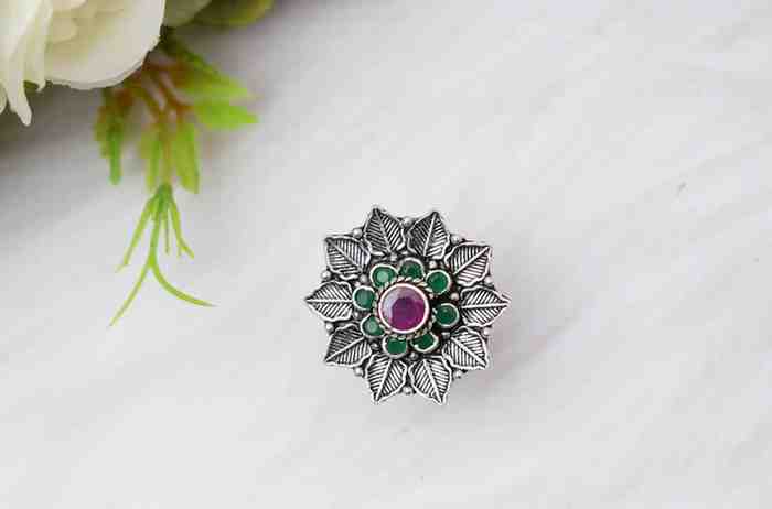 A silver ring featuring a floral design with a red cutstone surrounded by green cutstones.