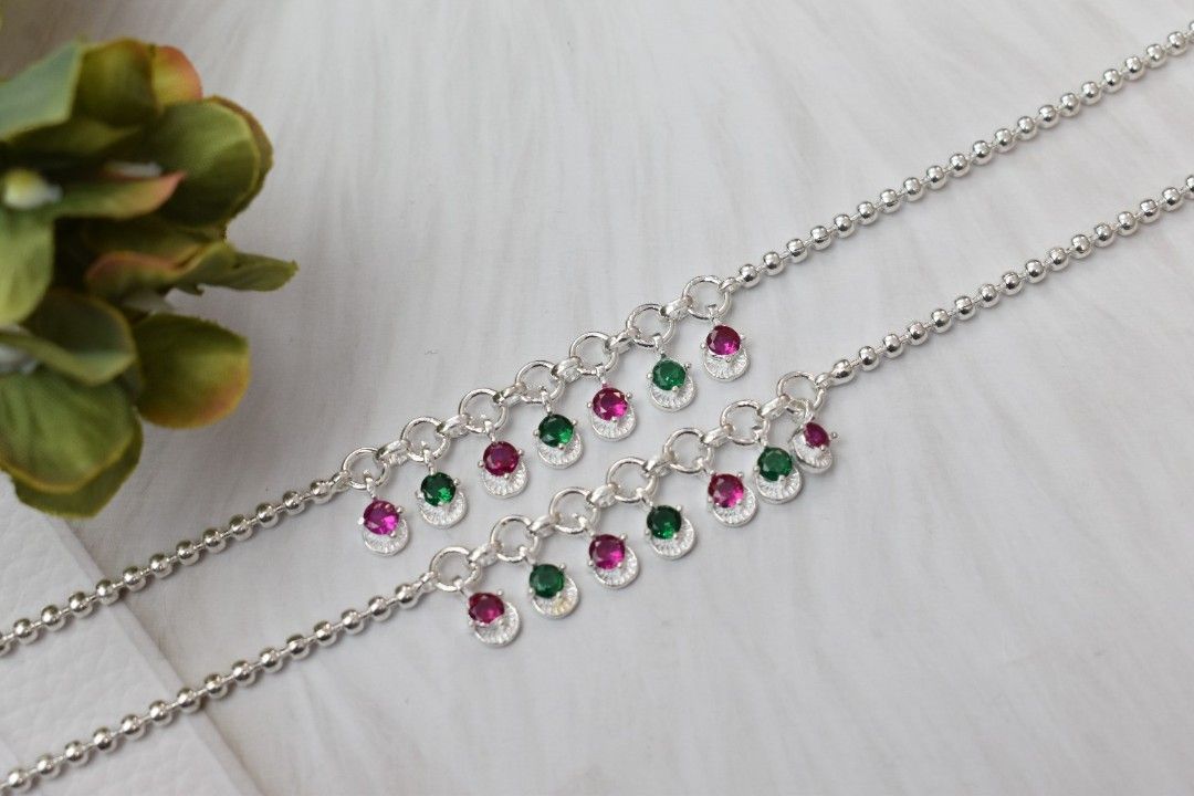 A pair of silver anklets with green and pink cutstones hanging from chains.