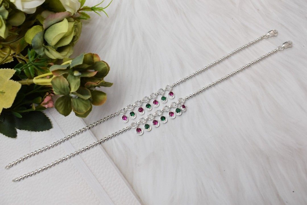 A pair of silver anklets with green and pink cutstones hanging from chains.