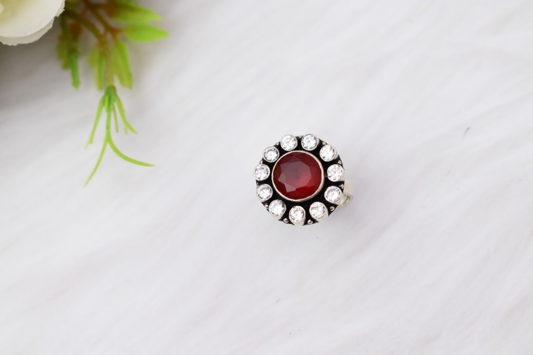 A silver ring with a circular design featuring a red cutstone surrounded by white cutstones.