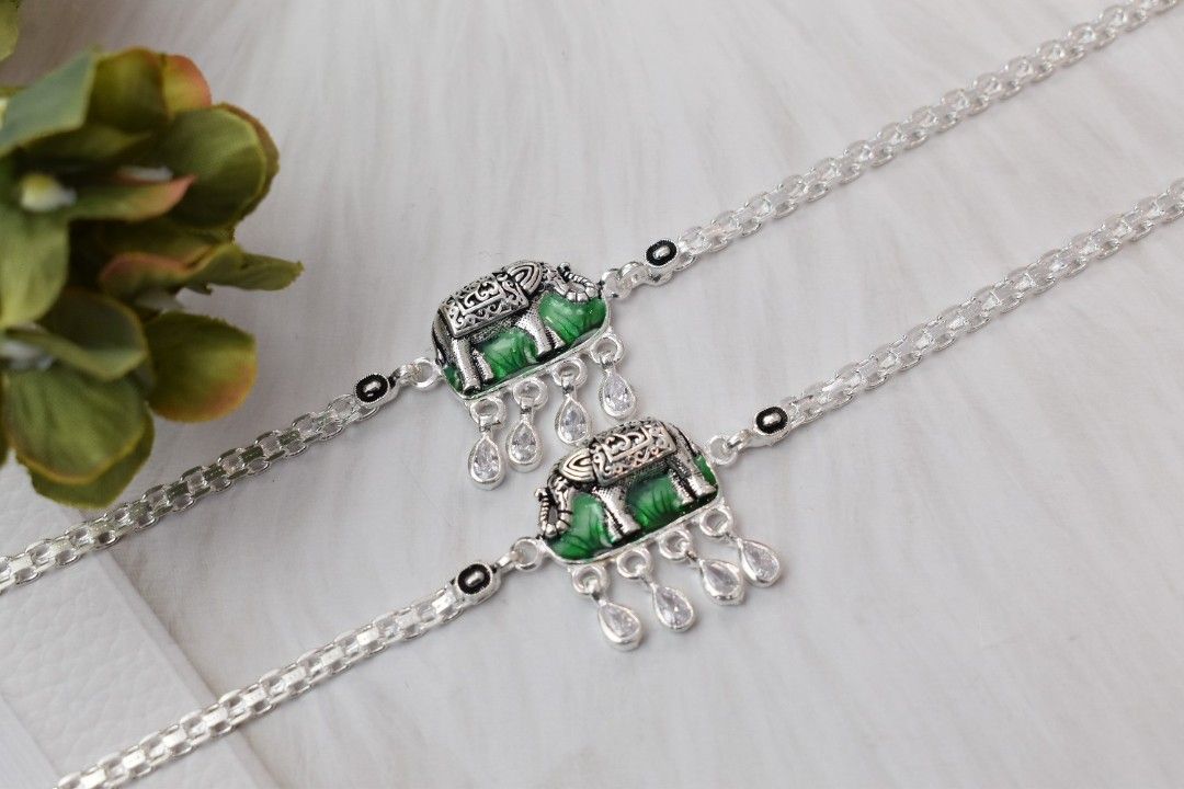 A pair of silver anklets with elephant-shaped pendants adorned with green enamel and white cutstones.