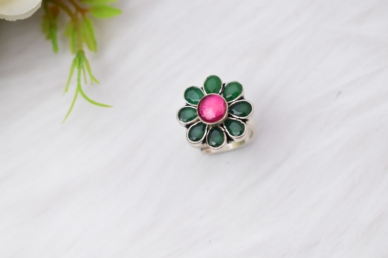 A silver ring with a floral design featuring a pink cutstone in the center and green cutstones surrounding it.