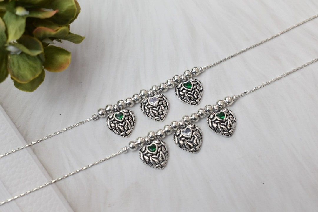 A pair of silver anklets with heart-shaped pendants adorned with green cutstones and beads.