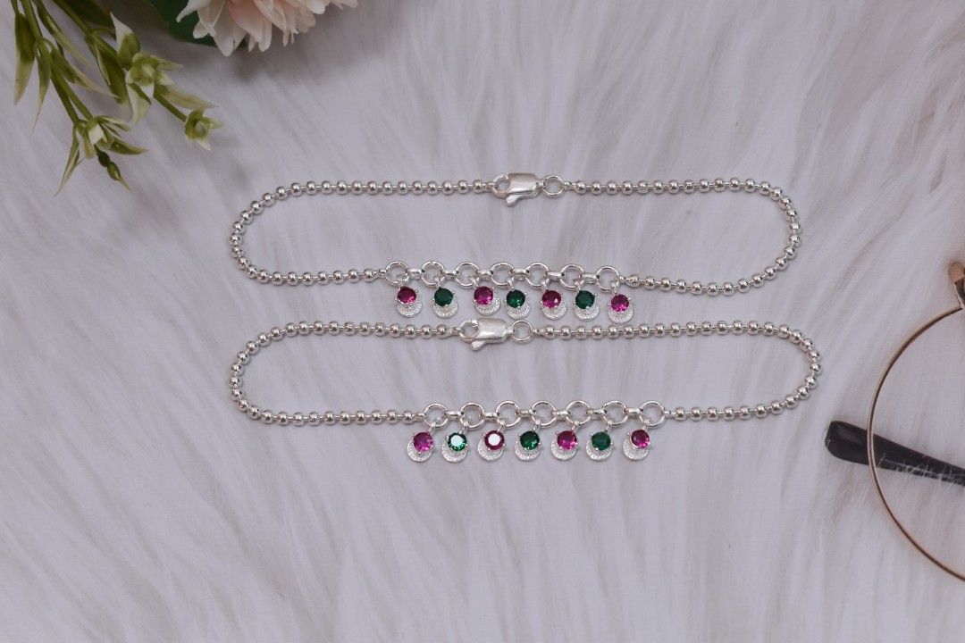 A pair of silver anklets with green and pink cutstones hanging from chains.