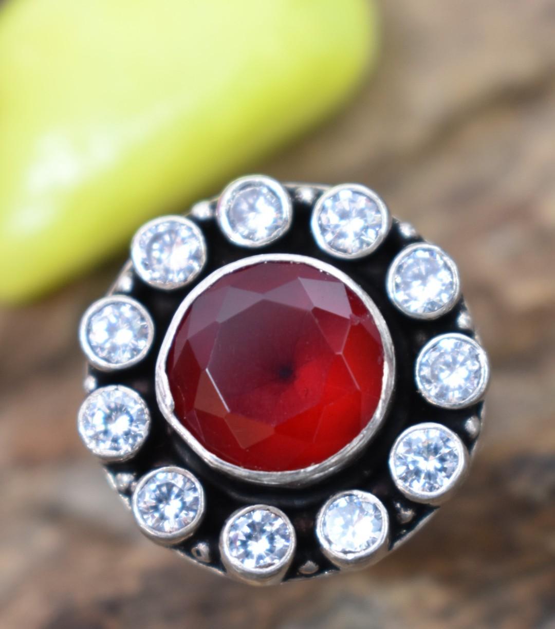 A silver ring with a circular design featuring a red cutstone surrounded by white cutstones.