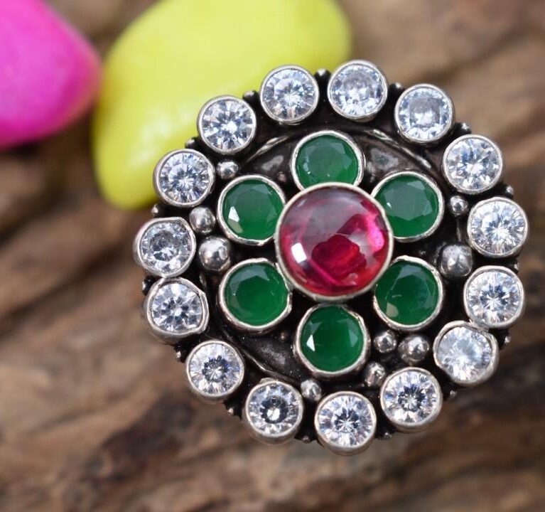 A silver ring with a circular design featuring a red cutstone surrounded by green and white cutstones.