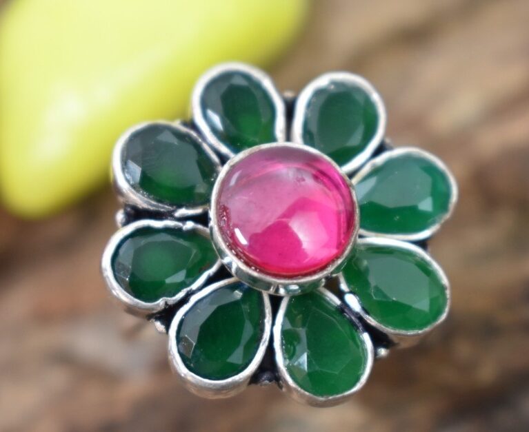 A silver ring with a floral design featuring a pink cutstone in the center and green cutstones surrounding it.