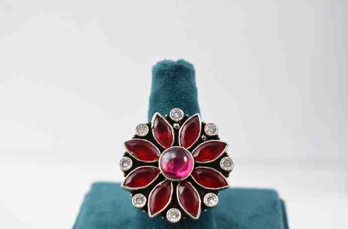 A silver ring with a floral design featuring red and pink cutstones surrounded by white cutstones.