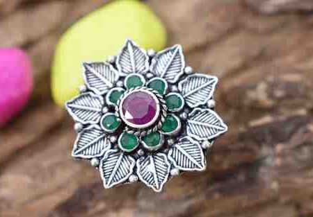 A silver ring featuring a floral design with a red cutstone surrounded by green cutstones.