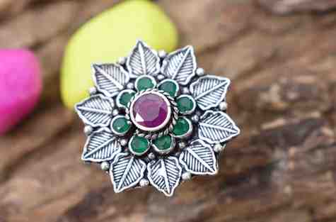 A silver ring featuring a floral design with a red cutstone surrounded by green cutstones.