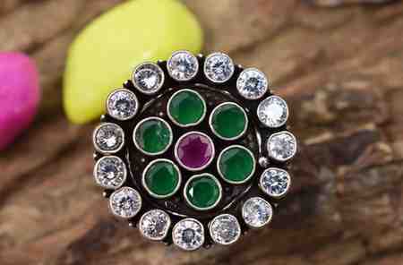 A silver ring with a circular design featuring a red cutstone surrounded by green and white cutstones.