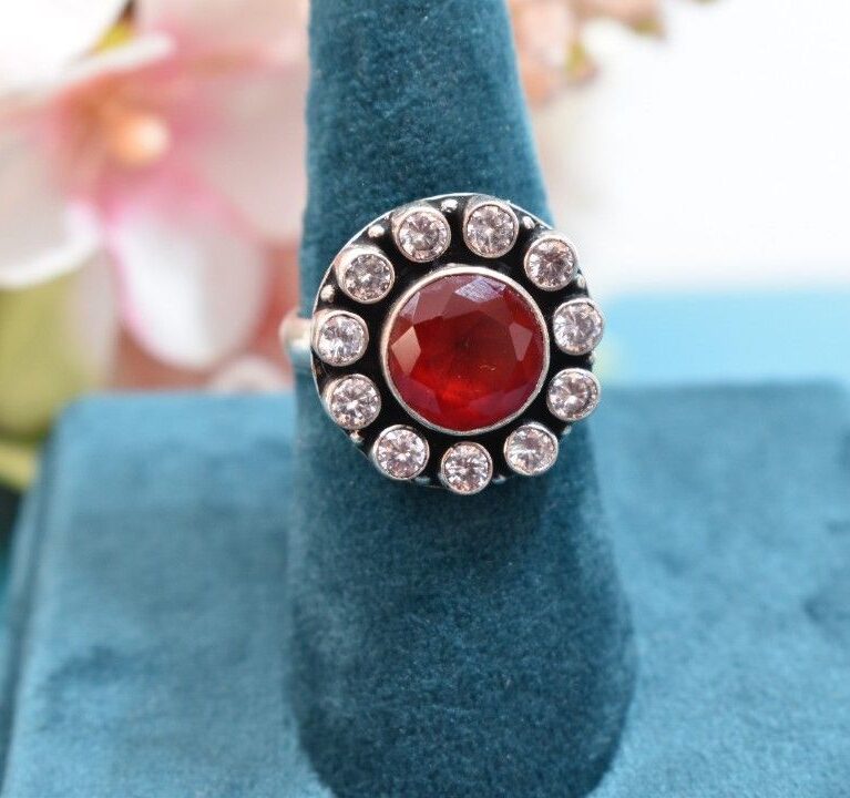 A silver ring with a circular design featuring a red cutstone surrounded by white cutstones.