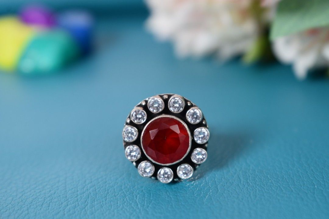 A silver ring with a circular design featuring a red cutstone surrounded by white cutstones.