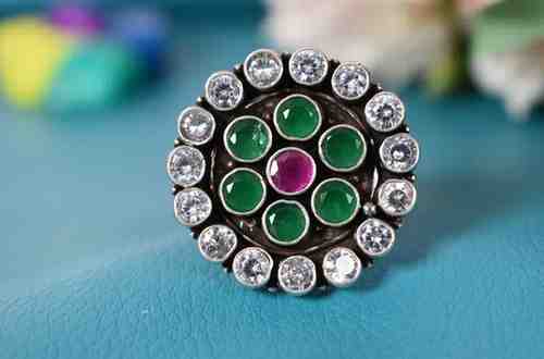 A silver ring with a circular design featuring a red cutstone surrounded by green and white cutstones.