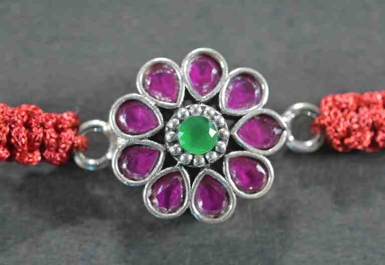 A silver Rakhi featuring a round red stone at the center surrounded by green stones.