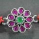 A silver Rakhi featuring a round red stone at the center surrounded by green stones.